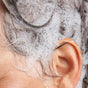 Close-up of a persons ear and wet hair covered in white NOTICE Hair Co The Detoxifier Shampoo foam, with bubbles and lather on the scalp and around the ear during a clarifying treatment.