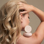 A blonde woman with wavy hair shows her profile, holding her hand to her head. Two NOTICE Hair Co The Fixer Conditioner bars rest on her bare shoulder, highlighting plastic-free hair care against a neutral background.