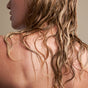 A close-up of damp, wavy blonde hair cascading down a person’s back and shoulder—showcasing results from NOTICE Hair Co’s The Fixer Conditioner. Skin and hair glisten against a neutral beige backdrop; the face is partially in profile.