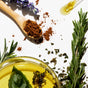 The Fixer Conditioner by NOTICE Hair Co is shown with a wooden spoon of brown powder, yellow oil, rosemary sprigs, and dried herbs—plastic-free essentials for repairing damaged hair on a white background.