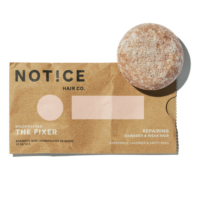 A round, brown Notice Hair Co The Fixer Shampoo bar rests on a labeled paper wrapper, offering a plastic-free solution to repair damaged and weak hair with a lavender and sweet basil scent.