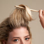 A person with blonde hair uses The Fixer Shampoo from NOTICE Hair Co, embracing plastic-free hair care while styling the front section of their hair against a neutral background.