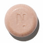 The Baby Cleanse Shampoo Bar - Sensitive & Dry Hair by NOTICE Hair Co is a round, light pink bar embossed with an "N" and dot, ideal for sensitive skin, shown casting a shadow on a white background.