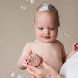 A baby with soap bubbles on their head looks at a round NOTICE Hair Co Baby Cleanse Shampoo Bar—marked with an N—held by an adult, as bubbles float around. Perfect for sensitive, dry hair and gentle, plastic-free care.