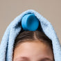 The NOTICE Hair Co Kids Conditioner Bar - For Tangles, in blue, sits atop a child's wet brown hair, partially wrapped in a light blue towel against a plain beige background.