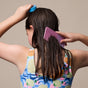 A child in a colorful swimsuit uses a blue sponge and wide-toothed pink comb to detangle wet brown hair—demonstrating the NOTICE Hair Co Kids Conditioner Bar - For Tangles, viewed from behind against a plain background.