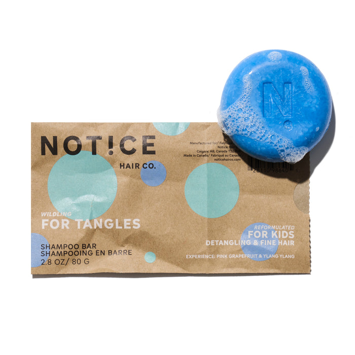 A blue Kids Shampoo Bar - For Tangles from NOTICE Hair Co sits on brown and blue packaging, designed for detangling and fine hair, with suds visible.