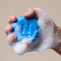 A hand grips the NOTICE Hair Co Kids Shampoo Bar - For Tangles, blue with an embossed N and thick white foam, held against a light background—ideal for gentle detangling and caring for fine hair.