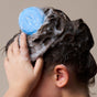 A person lathers the NOTICE Hair Co Kids Shampoo Bar - For Tangles into their wet, dark hair, creating foamy suds. The focus is on the hand, hair, and shampoo bar against a neutral background.