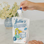 A hand pours a scoop of Nellie's Oxygen Brightener into an open bag on a white countertop, with a vase of daisies in the background.