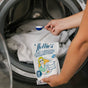 A person adds a scoop of Nellie’s Oxygen Brightener to a washing machine of whites, holding the Oxygen Brightener packaging in front of the open washer.