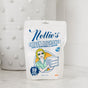 A white pouch of Nellie's Oxygen Brightener laundry powder rests on a marble countertop. The retro package features a cartoon woman with towels and highlights it as a hypoallergenic bleach alternative with 50 scoops (800g/1.77 lb).