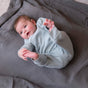 A baby in a light blue onesie lies on a gray Naturepedic Organic Breathable Swaddle Blanket from the Multi-Color Pack, reaching for their feet with both hands and gazing upward with curiosity.