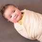 A baby wrapped in a Naturepedic Organic Breathable Swaddle Blanket from the Multi-Color Pack lies on a textured gray surface, gazing upward with rosy cheeks and a gentle smile.