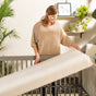 A woman smiles as she places a Naturepedic Classic Organic Cotton Baby Crib Mattress into a gray crib in a bright, cozy room with plants and light-colored decor.