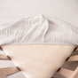 A close-up of a bed corner shows a Naturepedic Organic Breathable Muslin Crib Sheet partially pulled back on a light wooden frame, revealing a beige mattress protector beneath for breathable comfort and softness.