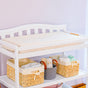 A Naturepedic Organic Cotton Changing Pad sits atop a white baby changing table (16.5 x 31.5 x 4 in) against a light purple wall. The lower shelf holds two wicker baskets and one gray basket stocked with baby supplies.