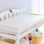 A Naturepedic Organic Cotton Changing Pad with safety straps is placed by a window atop a white table (16.5 x 31.5 x 4). Baskets below hold baby items, and soft natural light fills the room.