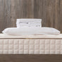 A neatly folded stack of Naturepedic Percale Sheet Set - GOTS Certified Organic Cotton and a pillowcase rest on a beige quilted mattress with a wooden wall background, evoking luxury hotel bedding.