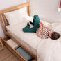 A young child in striped pajamas lies on a neatly made bed with the Naturepedic Percale Sheet Set (GOTS Certified Organic Cotton), holding a toy. The wooden bed’s partially open storage drawer reveals folded blankets inside.