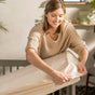 A woman smiles while putting a Naturepedic Fitted Waterproof Crib Mattress Protector Pad on a mattress in a softly lit room. She wears a beige sweater, and the background features decor and greenery on a shelf.