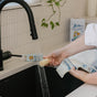 Someone holds a denim garment and Nellie's WOW Stick Stain Remover under water from a black kitchen faucet, with green cleaning products and a potted plant on the counter.
