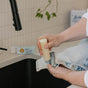Someone rubs Nellie's WOW Stick Stain Remover on the cuff of light-wash denim jeans over a kitchen sink, with a plant and other green cleaning products visible in the background.