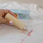 A hand applies Nellie's WOW Stick Stain Remover to a stained white shirt. In the background, a blue box labeled Nellie's WOW Stick highlights this natural stain remover, ideal for eco-friendly cleaning.