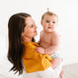 A smiling woman in a white shirt holds her baby, with a Natemia Organic Super Absorbent Washcloth made from 100% GOTS Certified Organic Cotton draped over her shoulder. The baby is in a diaper against a bright, simple background.