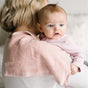 A baby with light brown hair in pink is held over an adult’s shoulder, resting on a Natemia Organic Super Absorbent Washcloth made from 100% GOTS Certified Organic Cotton. The baby looks alert and gazes off to the side.