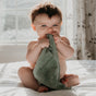 A baby with tousled brown hair sits on a sunlit bed, chewing on a green Natemia Organic Super Absorbent Washcloth made from 100% GOTS Certified Organic Cotton, with a soft, bright window in the background.