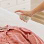 A hand sprays Nellies Wrinkle-B-Gone - Lemongrass onto a pink button-up shirt on a bed, with a white dresser and softly lit bedroom in the background, highlighting an easy, green wrinkle-removing routine.