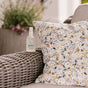 A bottle of Nellies Wrinkle-B-Gone - Lemongrass sits on a floral pillow atop a wicker chair, with blurred greenery and purple flowers in the background.