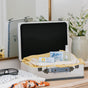 An open suitcase on a dresser holds folded yellow and white clothes, Nellies cleaning products, Wrinkle-B-Gone - Lemongrass, and a fabric refresher. Nearby are a vase with white flowers, sunglasses, extra clothes, and a mirror reflecting the scene.