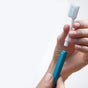 A person holds a white Nada Soft Adult Toothbrush head in one hand and a blue handle in the other, preparing to attach it—an easy, eco-friendly way to reduce waste by reusing the toothbrush handle.