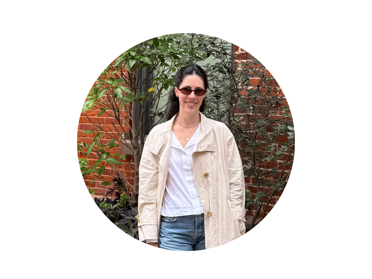 A woman with long dark hair, wearing sunglasses, a light-colored coat, white shirt, and blue jeans, stands outdoors in front of green foliage and a brick wall.