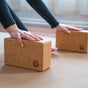 Close-up of hands pressing on a Yoloha Yoga Cork Yoga Block from the Cork Yoga Block Set, positioned on a mat. The person, dressed in long sleeves, is practicing an indoor yoga pose, possibly referencing the included yoga block guidebook for guidance.