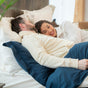 A couple cuddles under a blue blanket, smiling in bed with white pillows and a Naturepedic Percale Sheet Set made from GOTS Certified Organic Cotton, surrounded by luxury bedding in a bright, cozy bedroom.