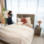 A woman playfully raises a pillow as if to hit a smiling man sitting up in bed. The bright room showcases plants, bedside tables, and Naturepedic’s GOTS Certified Organic Cotton Percale Sheet Set, adding elegance to the inviting scene.