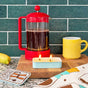 A lit The Neighborgoods Soy Wax Candle in a rectangular sardine tin sits on a wooden board next to a red French press, a yellow mug, and a banana.