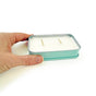 A hand holds a rectangular teal metal sardine tin containing a The Neighborgoods Soy Wax Candle with two wicks.
