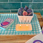 A The Neighborgoods Soy Wax Candle with two lit wicks sits on a blue and white checkered tray next to a ceramic basket filled with fresh figs and a closed sardine tin candle.