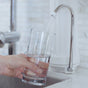 A hand fills a clear glass with water from the New Wave Enviro 10-Stage Countertop Water Filter in a bright kitchen.