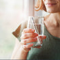 A woman stands by a sunlit window, holding a glass of water with the New Wave Enviro Alkaline Water Wand inside, possibly raising pH as she enjoys the gentle natural light.