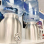 Two New Wave Enviro Stainless Steel Water Crocks with shiny metal bases and clear bottles are side by side on a white shelf, with framed pictures and colorful string lights in the background.