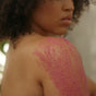 A person with curly hair applies NOPALERA's Cactus Body Polish, a pink exfoliant, to their bare shoulder. The focus is on the textured scrub and smooth skin, highlighting the revitalizing effects of this body polish.