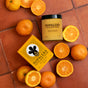 A jar and box of NOPALERA Cactus Body Polish are displayed on terracotta tiles, surrounded by whole and halved oranges. The bright yellow packaging with bold black text and design stands out against the vibrant setting.