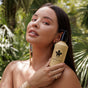 A woman with long dark hair holds a NOPALERA Cactus Body Cream - Dulce De Cuerpo near her face, standing outdoors among lush tropical plants. She wears earrings, a ring, and a gold cuff bracelet.