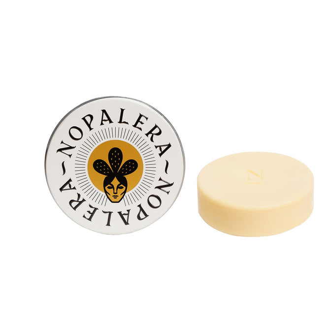 A round tin labeled “NOPALERA” with a stylized face and prickly pear design sits beside a plastic-free, light yellow Moisturizing Botanical Bar embossed with the letter “N.”.