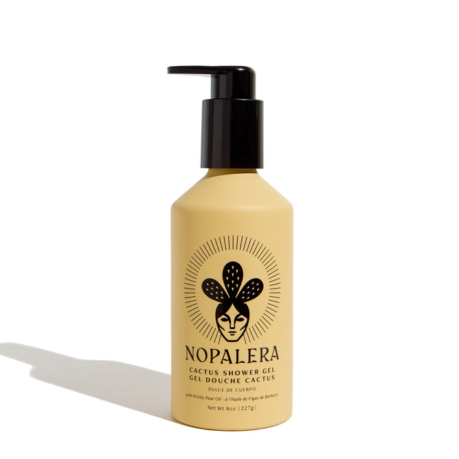 A beige NOPALERA Cactus Shower Gel bottle with a black pump, stylized face and cactus logo. The vegan gel features prickly pear oil; label text appears in English and French. The product casts a shadow to the left.